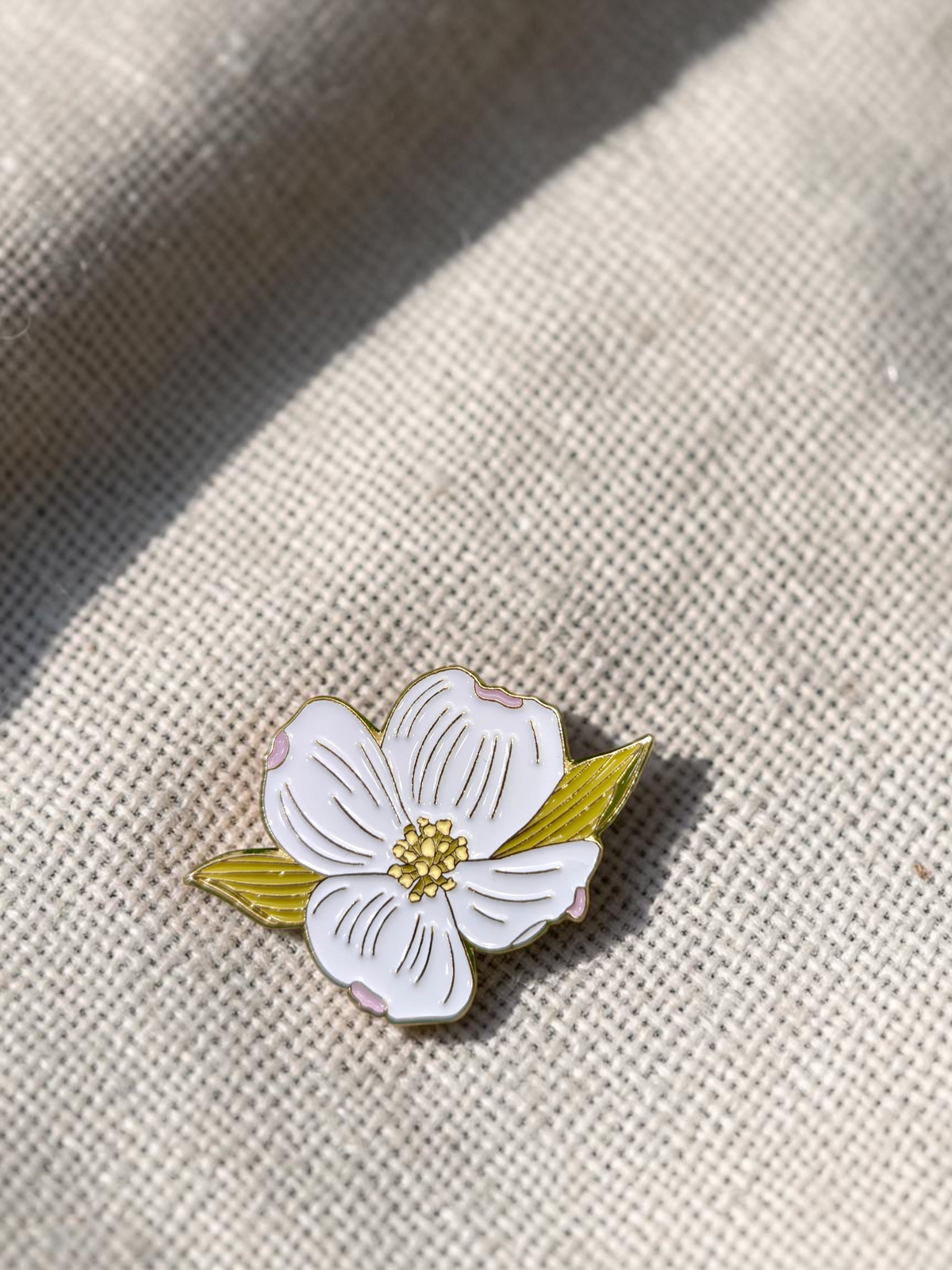The Plant Ecologist flowering dogwood enamel pin with white petals and a golden center resting on a green leaf.
